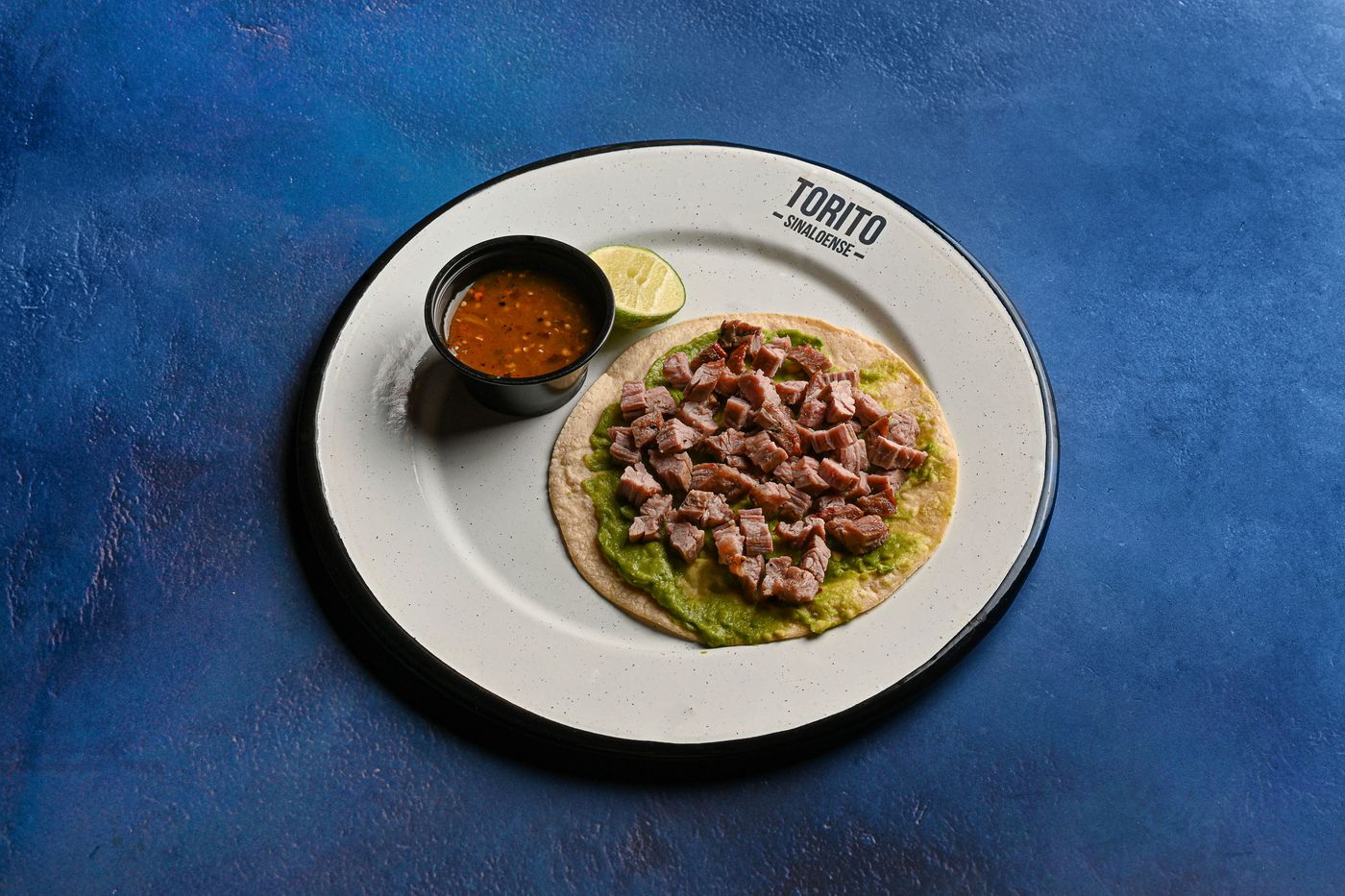 A single beef taco beside a cup of consommé on a white plate