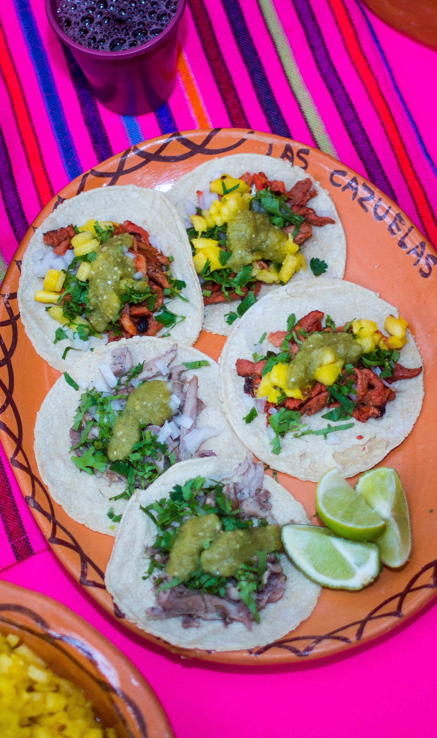 A row of soft tacos topped with cilantro and onion on a board with a cup of red salsa and lime