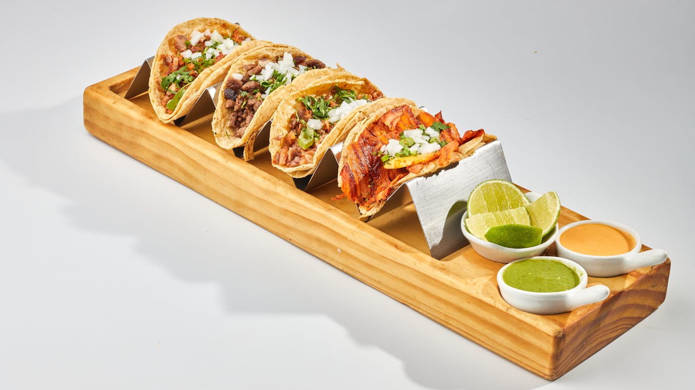 A wooden tray of tacos with lime wedges and dipping sauces