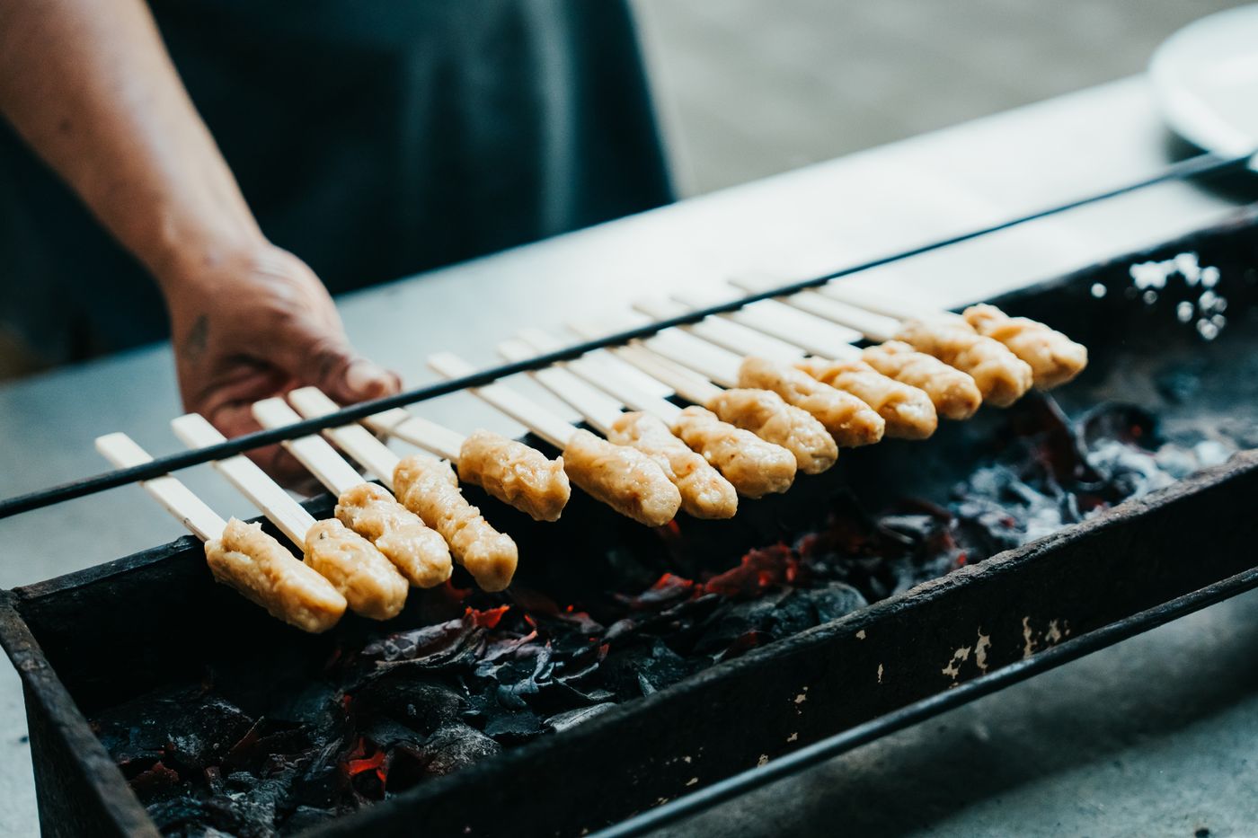 A cook's hand lays a long row of pale chicken skewers across a charcoal grill, red embers glowing in the trough below