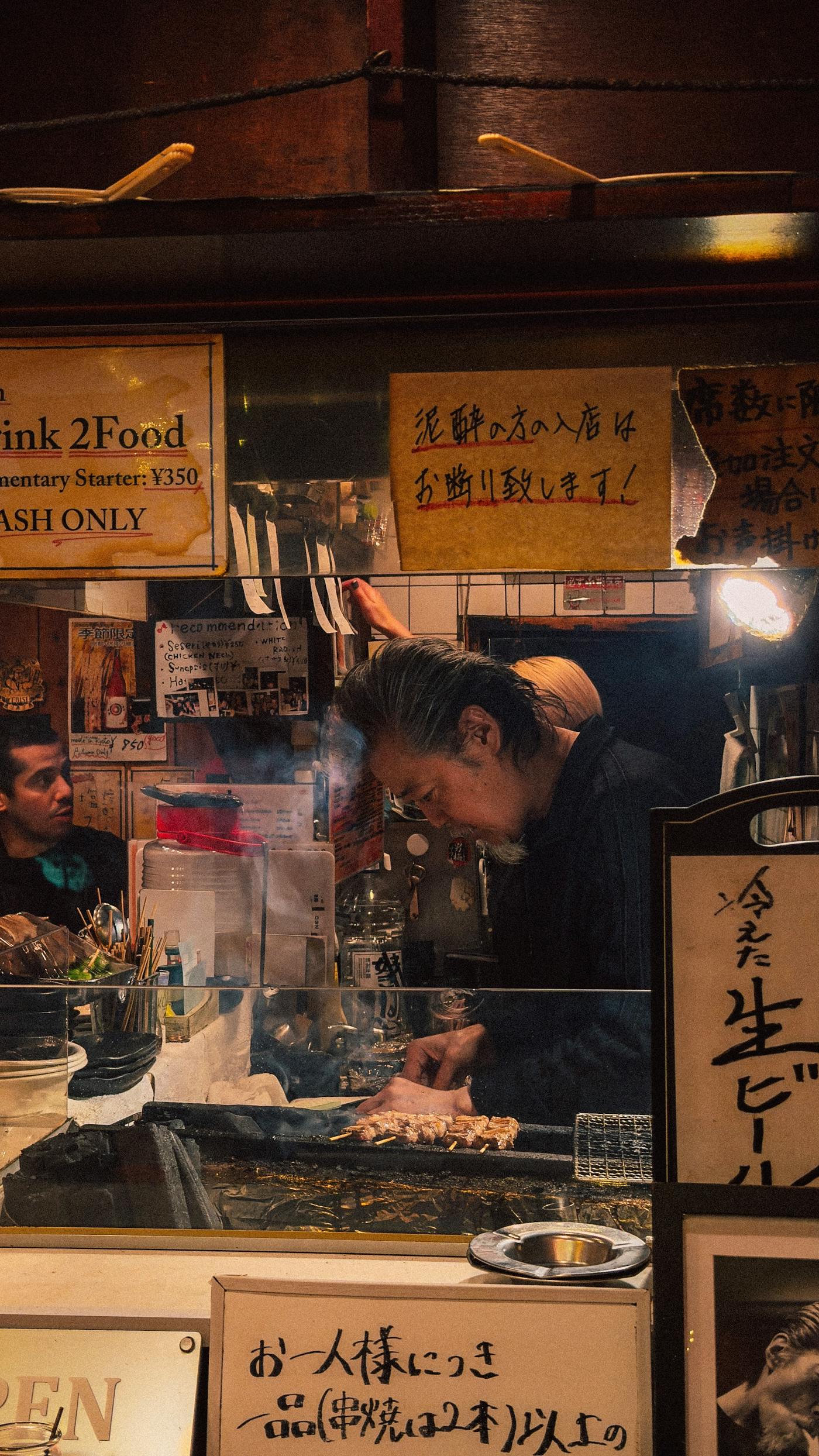 A grill cook leans over the counter of a small lantern-lit izakaya at night, tending skewers above the coals