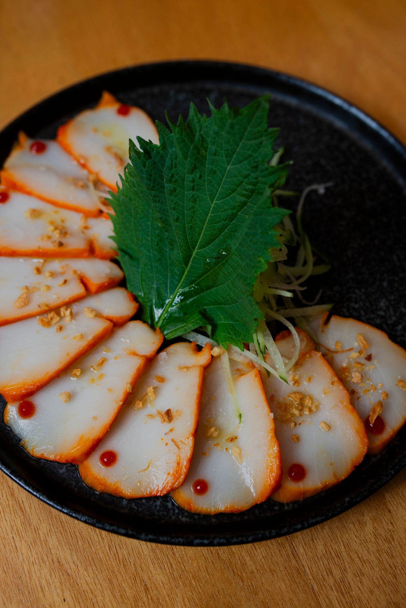 Seared white fish fanned across a black plate with a green shiso leaf, dressed with chili oil and crisp garlic