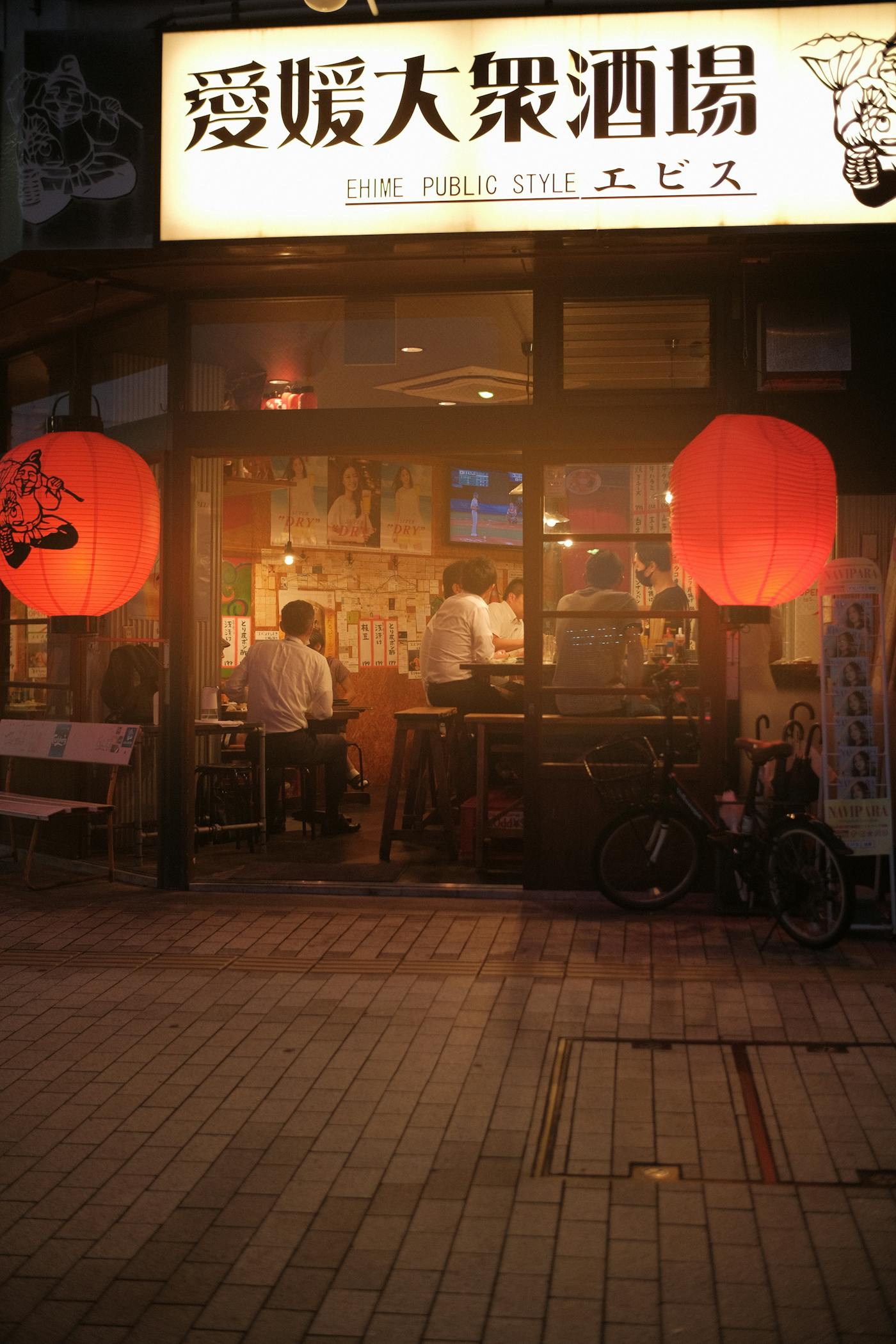 An izakaya storefront at night with two glowing red paper lanterns flanking the doorway and guests at the counter inside